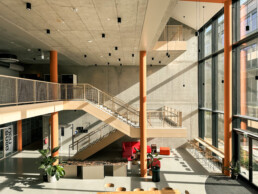 Zweist&ouml;ckige Hallee mit Betonwand, Treppe und orangenen Pfeilern. Auf der rechten Seite: eine Glasfront, die den Raum in warmes Licht taucht. In der Halle stehen M&ouml;bel &ndash; St&uuml;hle, ein Sofas, Pflanzen und eine Reihe Barhocker mit Tresen an der Glaswand